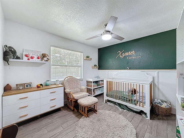 bedroom featuring wood finished floors, a nursery area, a textured ceiling, and a ceiling fan