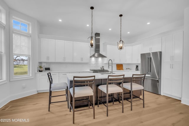 kitchen featuring wall chimney range hood, high end refrigerator, an island with sink, decorative light fixtures, and white cabinets