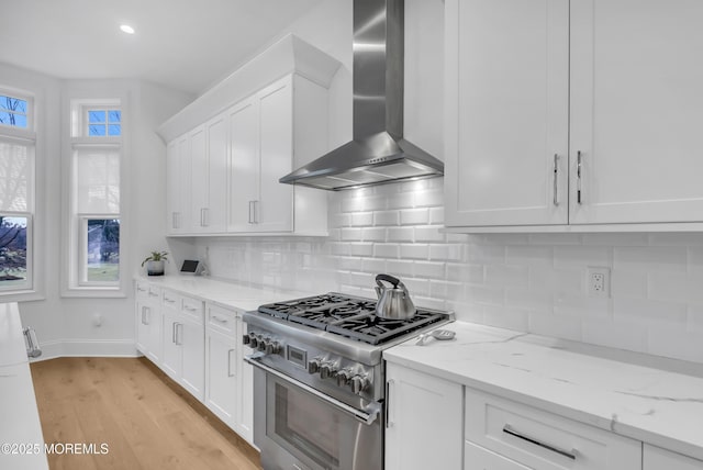 kitchen with wall chimney range hood, stainless steel stove, light stone countertops, light wood-type flooring, and white cabinetry