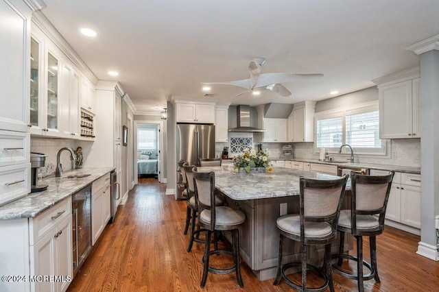 kitchen featuring wall chimney range hood, a kitchen breakfast bar, freestanding refrigerator, white cabinetry, and a sink