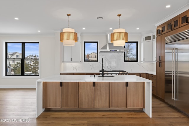 kitchen featuring an island with sink, stainless steel built in fridge, wall chimney range hood, and decorative light fixtures