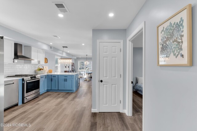 kitchen featuring gas stove, blue cabinetry, hanging light fixtures, wall chimney range hood, and kitchen peninsula