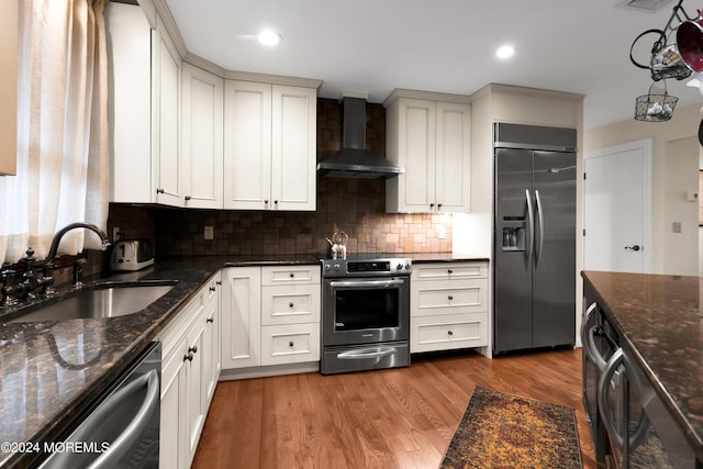 kitchen featuring wall chimney range hood, appliances with stainless steel finishes, hardwood / wood-style flooring, dark stone countertops, and sink