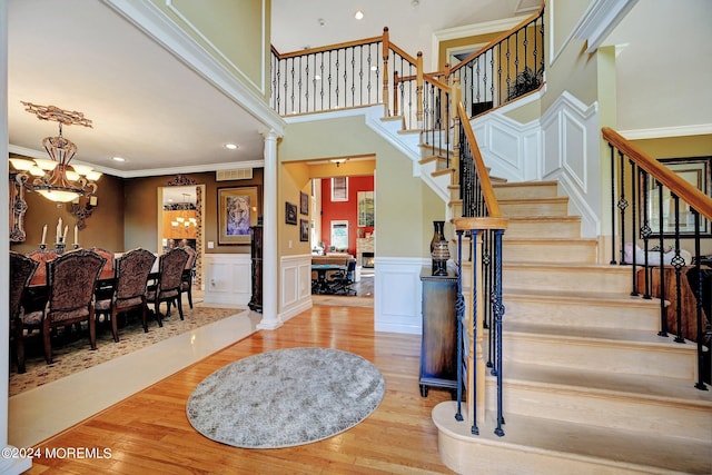 staircase with a wainscoted wall, wood finished floors, ornamental molding, an inviting chandelier, and ornate columns