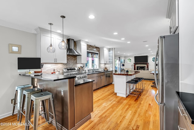 kitchen with pendant lighting, a kitchen breakfast bar, wall chimney exhaust hood, appliances with stainless steel finishes, and light hardwood / wood-style floors