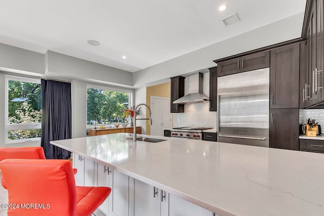 kitchen featuring wall chimney exhaust hood, a kitchen bar, stainless steel appliances, sink, and light stone counters