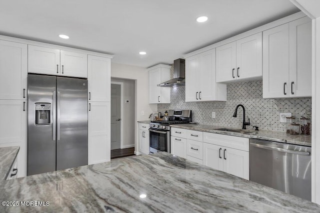 kitchen with light stone counters, appliances with stainless steel finishes, white cabinets, a sink, and wall chimney range hood