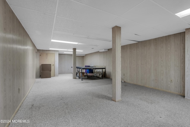 basement featuring light carpet, a drop ceiling, and wood walls