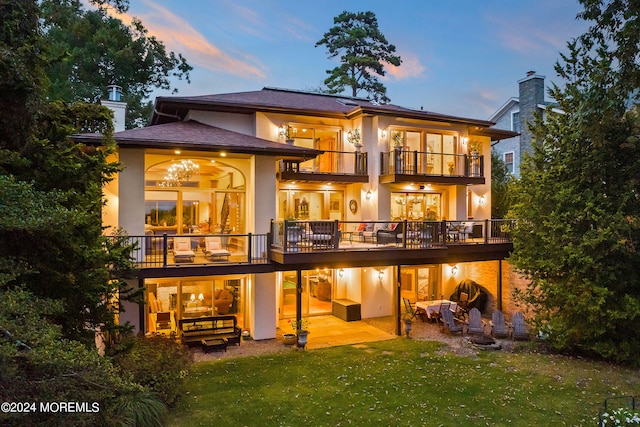 back house at dusk featuring a balcony, a lawn, and a patio