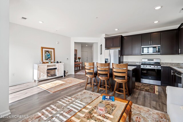 kitchen with dark brown cabinets, hardwood / wood-style flooring, appliances with stainless steel finishes, a breakfast bar, and light stone countertops