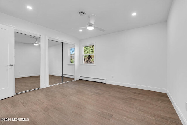 unfurnished bedroom featuring baseboard heating, ceiling fan, hardwood / wood-style floors, and two closets