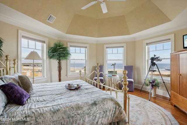 bedroom featuring a tray ceiling, ornamental molding, hardwood / wood-style floors, a water view, and ceiling fan