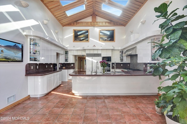 kitchen featuring a skylight, white cabinetry, wall chimney exhaust hood, high vaulted ceiling, and decorative backsplash