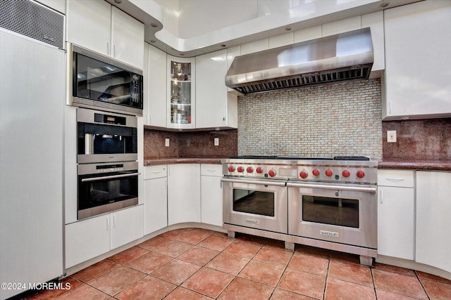 kitchen with wall chimney range hood, built in appliances, backsplash, light tile patterned flooring, and white cabinetry