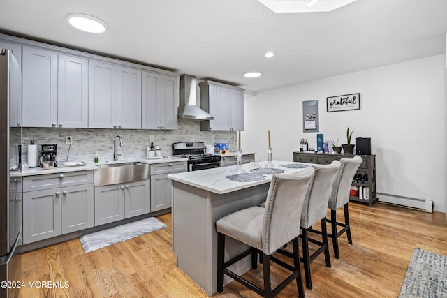 kitchen with wall chimney range hood, sink, a baseboard radiator, stainless steel gas range oven, and a center island with sink