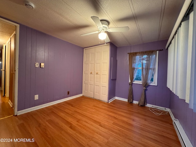 unfurnished bedroom with a textured ceiling, ceiling fan, wood-type flooring, a baseboard radiator, and wood walls