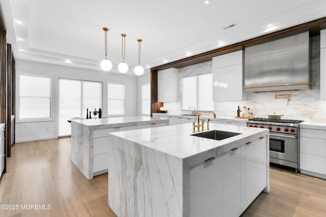 kitchen with white cabinets, wall chimney exhaust hood, an island with sink, and designer stove