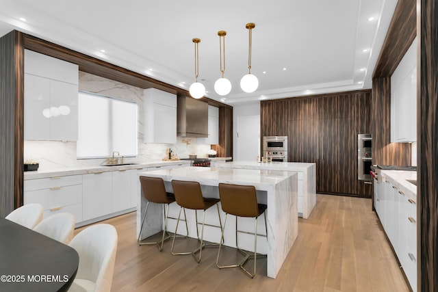 kitchen with decorative backsplash, wall chimney exhaust hood, sink, a large island with sink, and light hardwood / wood-style flooring