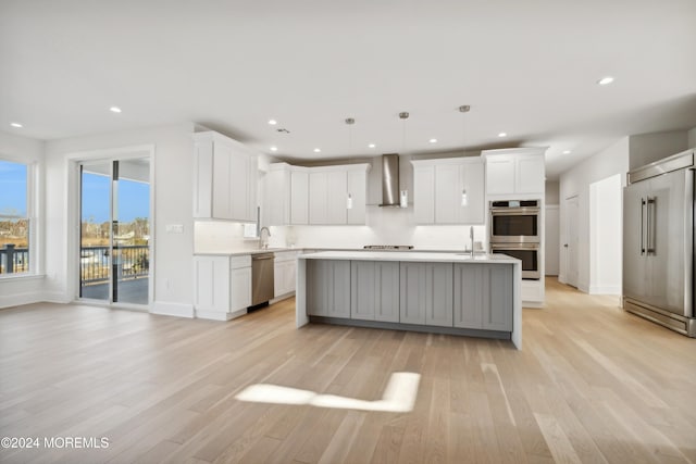 kitchen with stainless steel appliances, a kitchen island with sink, wall chimney range hood, decorative light fixtures, and white cabinetry