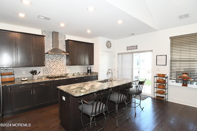 kitchen with wall chimney range hood, dark hardwood / wood-style flooring, light stone countertops, a breakfast bar, and a kitchen island with sink