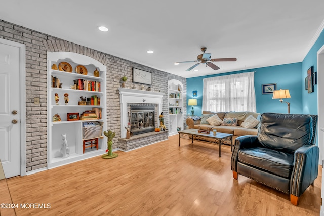 living room with built in shelves, ceiling fan, a brick fireplace, brick wall, and light wood-type flooring