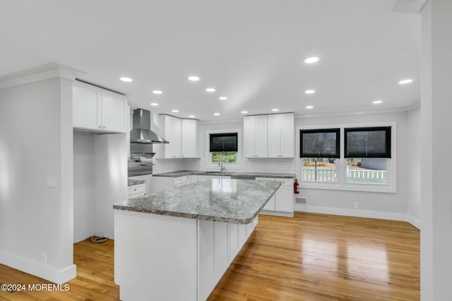 kitchen featuring a center island, wall chimney range hood, crown molding, light hardwood / wood-style flooring, and white cabinetry