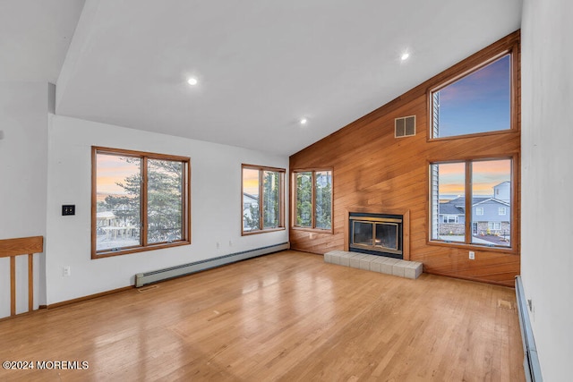 unfurnished living room with a baseboard radiator, light hardwood / wood-style flooring, high vaulted ceiling, wood walls, and a tiled fireplace