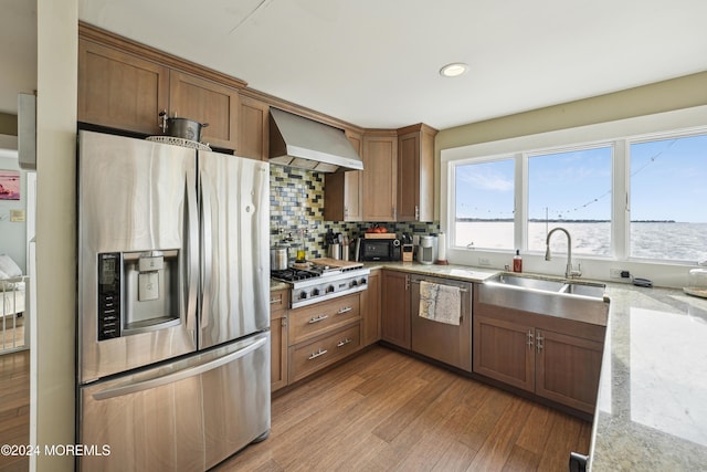 kitchen with sink, wall chimney exhaust hood, stainless steel appliances, light stone counters, and hardwood / wood-style flooring