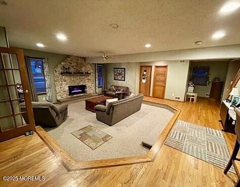 living room featuring a stone fireplace, ceiling fan, a textured ceiling, and light wood-type flooring