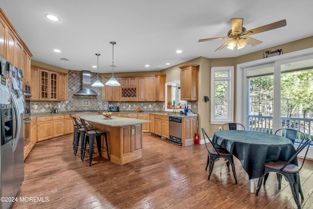 kitchen with ceiling fan, wall chimney exhaust hood, pendant lighting, a kitchen island, and appliances with stainless steel finishes