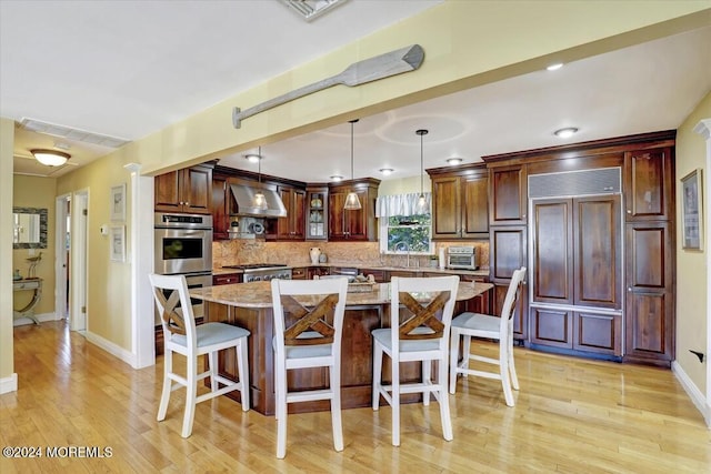 kitchen featuring a center island, wall chimney range hood, light stone countertops, decorative light fixtures, and stainless steel double oven