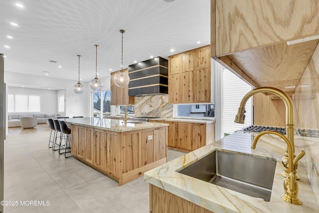 kitchen with light stone counters, backsplash, an island with sink, pendant lighting, and custom range hood