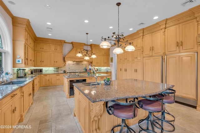 kitchen with light stone countertops, custom range hood, stainless steel appliances, sink, and a center island with sink