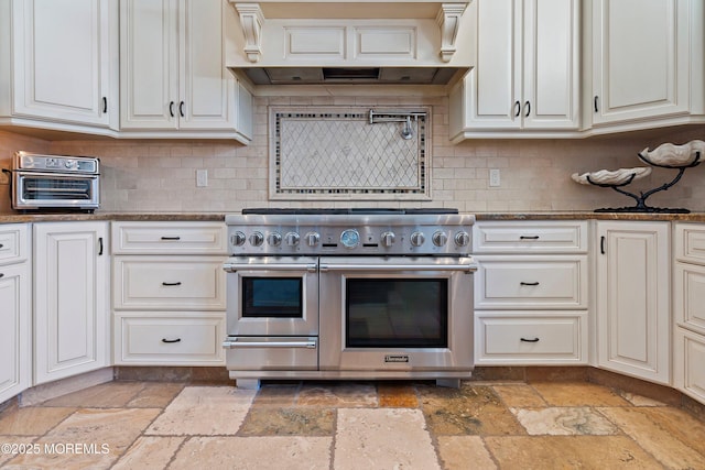 kitchen featuring decorative backsplash, premium range hood, double oven range, dark stone countertops, and white cabinets