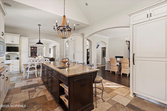 kitchen featuring pendant lighting, lofted ceiling, a kitchen island with sink, a notable chandelier, and light stone counters