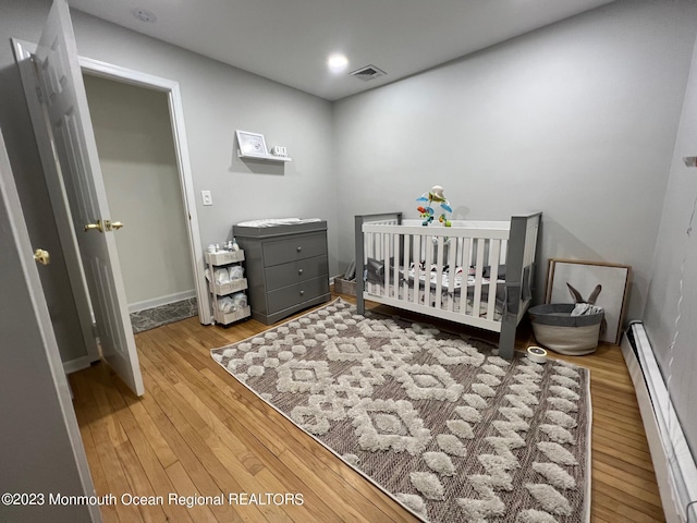 bedroom with a nursery area, wood-type flooring, and a baseboard heating unit