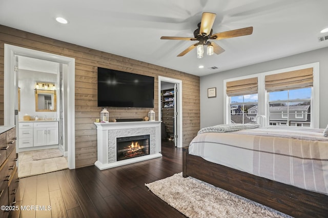 bedroom with wood walls, ensuite bathroom, a stone fireplace, ceiling fan, and a spacious closet