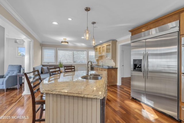 kitchen with a center island with sink, hanging light fixtures, built in refrigerator, light stone countertops, and sink