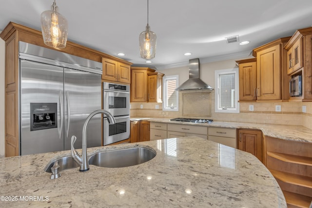 kitchen featuring light stone counters, wall chimney range hood, built in appliances, and decorative light fixtures