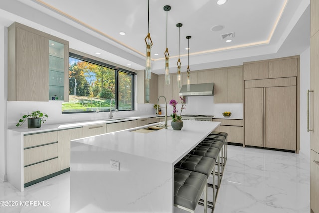 kitchen featuring light brown cabinetry, paneled refrigerator, a center island with sink, and a raised ceiling