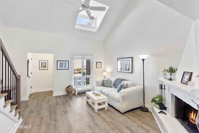 living room featuring a skylight, a brick fireplace, ceiling fan, high vaulted ceiling, and light hardwood / wood-style flooring