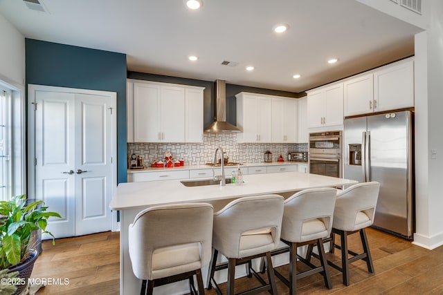 kitchen featuring a kitchen bar, stainless steel appliances, a kitchen island with sink, wall chimney range hood, and white cabinetry