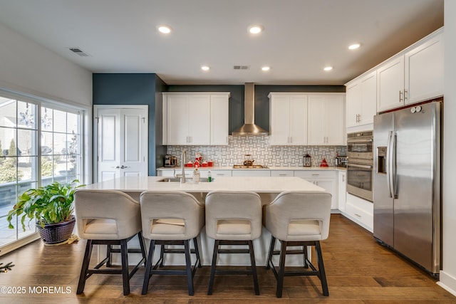 kitchen featuring stainless steel appliances, a kitchen island with sink, sink, wall chimney range hood, and white cabinetry