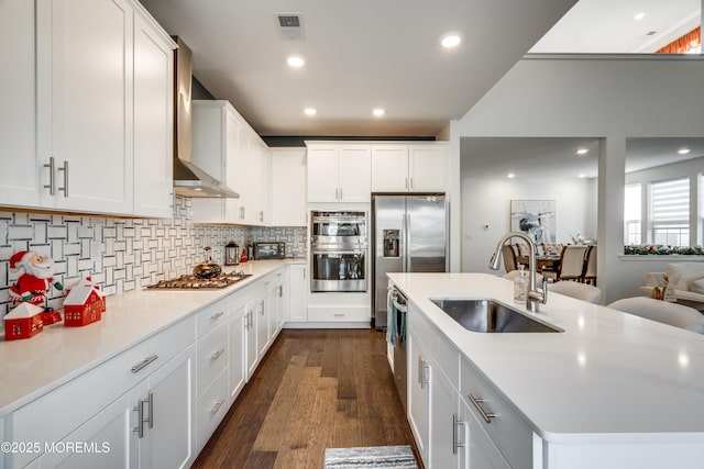 kitchen with white cabinetry, sink, wall chimney exhaust hood, stainless steel appliances, and an island with sink