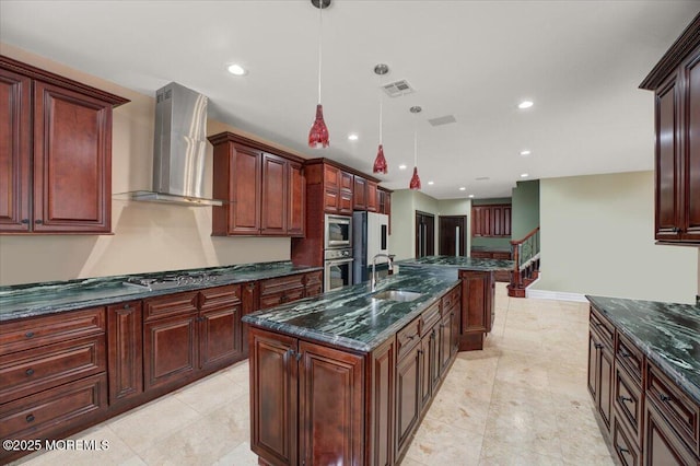 kitchen featuring sink, wall chimney exhaust hood, stainless steel appliances, decorative light fixtures, and a center island with sink