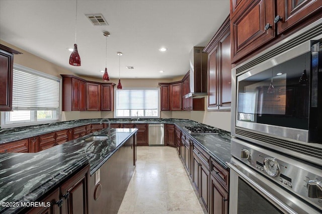 kitchen with appliances with stainless steel finishes, dark stone counters, wall chimney range hood, sink, and decorative light fixtures