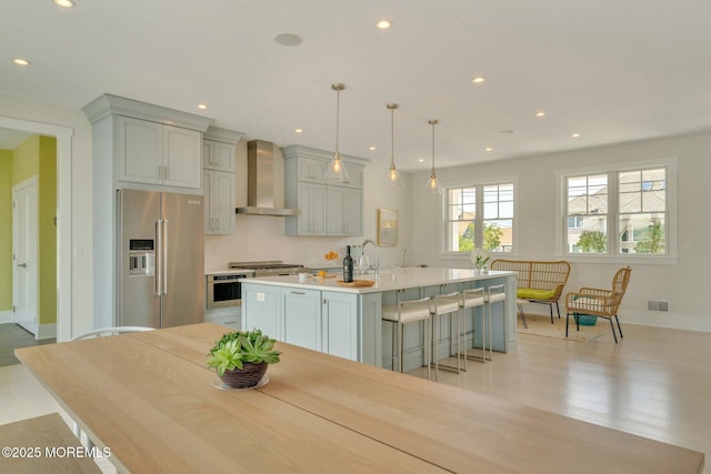 kitchen featuring appliances with stainless steel finishes, wall chimney exhaust hood, a kitchen island with sink, hanging light fixtures, and a breakfast bar area