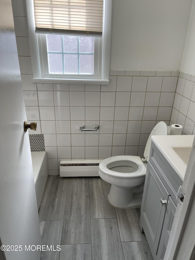 bathroom featuring a tub to relax in, toilet, tile walls, vanity, and a baseboard heating unit