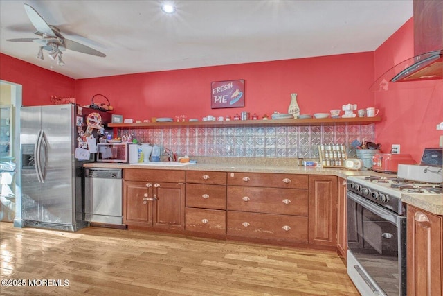 kitchen with appliances with stainless steel finishes, light wood-type flooring, ceiling fan, wall chimney exhaust hood, and sink