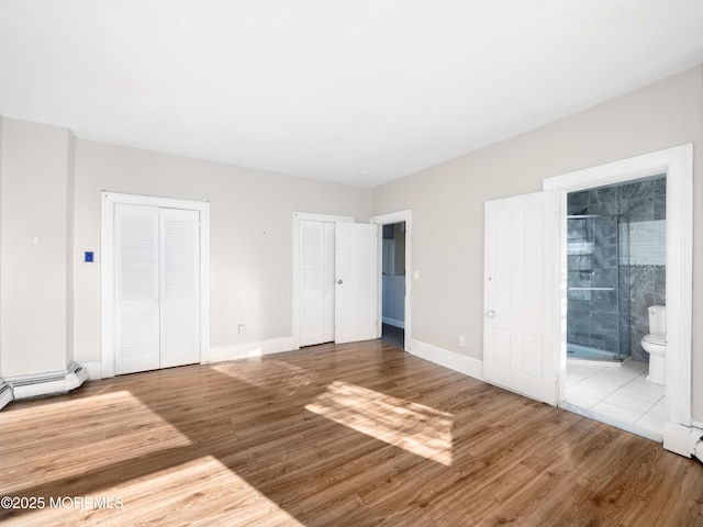 unfurnished bedroom with baseboard heating, two closets, light hardwood / wood-style flooring, and connected bathroom